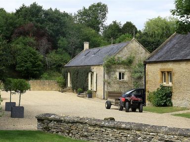 A gravel driveway with potted trees a stone wall a quad bike with a trailer and two stone buildings at The Granary in Bourton-On-The-Water