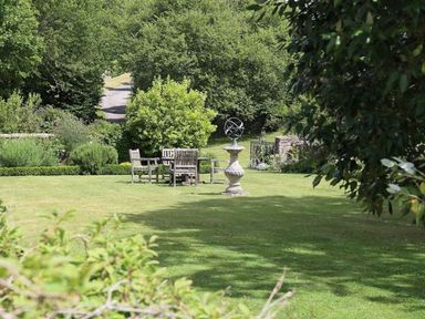 A garden with wooden chairs and table and a sundial on a lawn surrounded by trees at The Granary in Bourton-On-The-Water