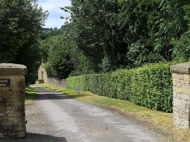 A gravel driveway lined with green hedges and trees leading to a stone building at The Granary in Bourton-On-The-Water