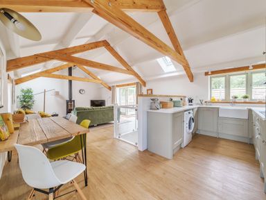 An open living space with wooden beams a dining table with chairs a green sofa a kitchen with appliances and windows at The Granary in Davidstow near Camelford