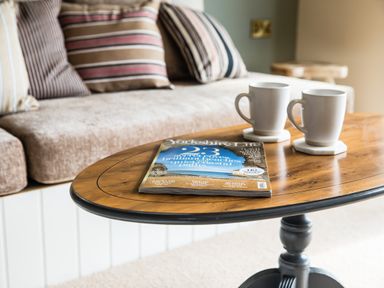 A wooden coffee table with two white mugs and a magazine in front of a sofa with striped cushions at Casa Formentera in Scarborough