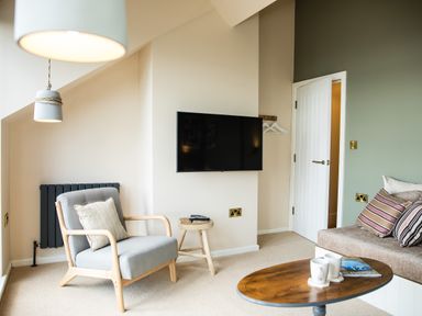 A living room with a gray armchair and pillow a wall-mounted television a wooden stool a round wooden coffee table with mugs and a magazine and a sofa with striped pillows at Casa Formentera in Scarborough