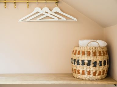 A wooden shelf with a woven basket containing towels and three white hangers on a brass rail on a peach wall at Casa Formentera in Scarborough