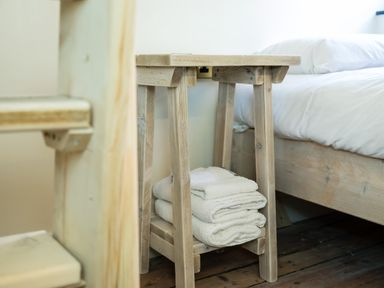 A small wooden side table with folded towels underneath next to a bed with white bedding