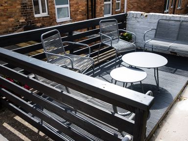An outdoor patio with metal chairs a bench and two round tables surrounded by a black wooden fence