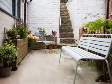 An outdoor patio area with a white bench, metal chair, various potted plants, and stone steps against a white brick wall at Casa Formentera in Scarborough