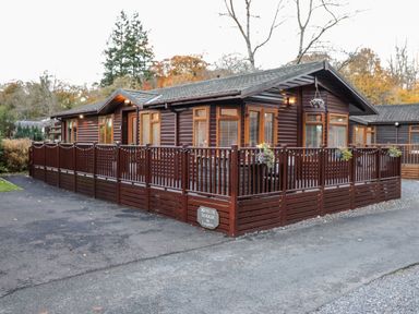 A wooden lodge with multiple windows and a fenced patio surrounded by trees at Ruskin Lodge Langdale 16 White Cross Bay near Windermere