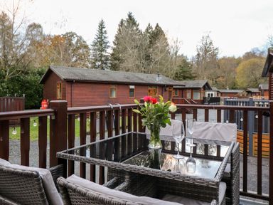 An outdoor patio with a glass table holding a vase of flowers and two wine glasses surrounded by wicker chairs and wooden cabins in the background at Ruskin Lodge in Langdale 16 White Cross Bay near Windermere