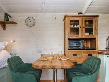 A small dining area with a wooden table two green chairs a wooden cabinet with glass doors a toaster oven and plates with croissants and glasses of orange juice at Lottie's Hut in Okehampton