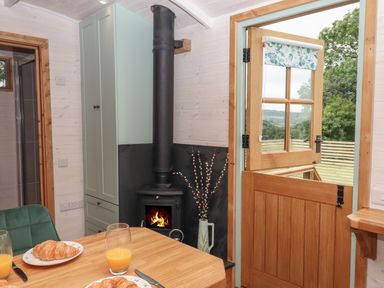 A dining area with a wooden table set with plates of croissants and glasses of orange juice a green chair a wood burning stove by a cabinet and a wooden door open to a view of trees and fields at Lotties Hut in Okehampton
