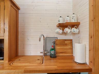 A kitchen countertop with a sink faucet cutting board paper towel holder and shelves with mugs and canisters at Lottie's Hut in Okehampton