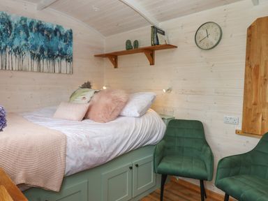 A bedroom with a bed with pillows a shelf with books and decorations a wall clock and two green chairs at Lottie's Hut in Okehampton