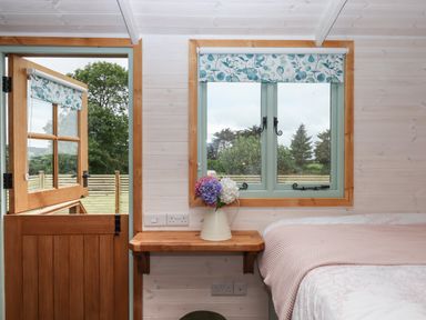 A wooden door and window with floral blinds next to a bed and a small wooden shelf with a vase of flowers at Lottie's Hut in Okehampton