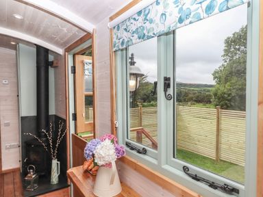 An interior view of a room with a wooden window overlooking a garden with a fence a jug with flowers and pastries on a wooden surface and a wood burning stove in the corner at Lottie's Hut in Okehampton