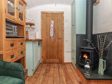 A kitchen area with wooden cabinets a green chair a wood stove with fire a wooden door at Lottie's Hut in Okehampton