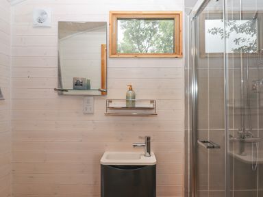 A bathroom with a small sink cabinet mirror glass shelf soap dispenser window and shower cubicle at Lottie's Hut in Okehampton