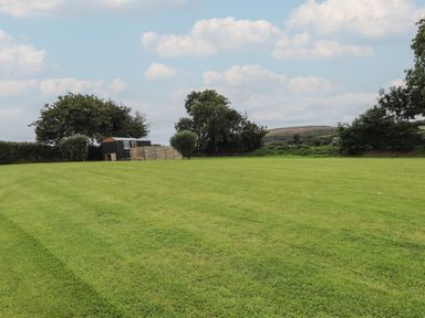 A grassy field with trees and a small cabin in the background at Lottie's Hut in Okehampton
