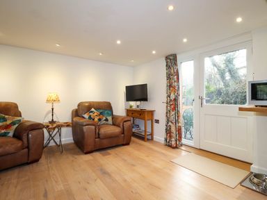 A living room with two leather armchairs a small wooden table a lamp a wall-mounted television a wooden console table with board games floral curtains and a glass door at The Studio at Dan y Garn in Newport Pembrokeshire