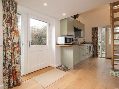 A kitchen area with green cabinets a microwave floral curtains and wooden stairs at The Studio at Dan y Garn in Newport Pembrokeshire