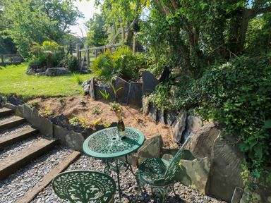 A green metal table and chairs set on gravel near steps beside a garden with plants and rocks at The Studio at Dan y Garn in Newport Pembrokeshire