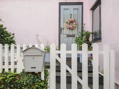 A front door with a floral wreath behind a white picket fence with a mailbox and plants at Moons Cottage in Fowey