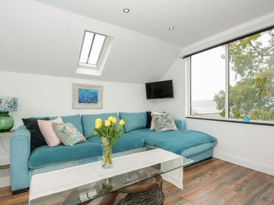 A living room with a blue sofa a glass coffee table with yellow flowers and a window showing trees at Moons Cottage in Fowey