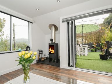 A room with a wood-burning stove a glass vase with yellow flowers on a table and a view of a garden with a table chairs and a green umbrella at Moon's Cottage in Fowey