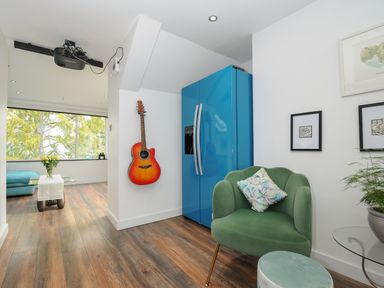 A room with a green armchair, blue refrigerator, hanging guitar, wooden floor, and a glass table with flowers at Moon's Cottage in Fowey