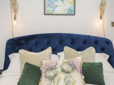 A bed with a blue upholstered headboard and multiple decorative pillows with hanging light bulbs on each side and a painting above the bed at Moon's Cottage in Fowey