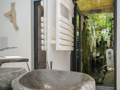 A stone sink with a faucet and a mirror above it next to a white towel radiator and an outdoor shower area with hanging white flowers and greenery at Moons Cottage in Fowey