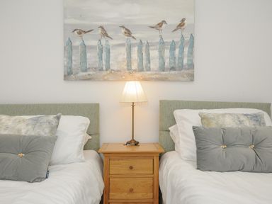 A bedroom with two single beds separated by a wooden nightstand with a lamp and a painting of birds on a fence above