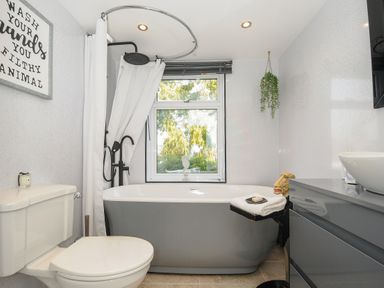 A bathroom with a toilet a gray bathtub with a shower curtain and black showerhead a sink with a gray cabinet and a window with a plant hanging from the ceiling at Moons Cottage in Fowey