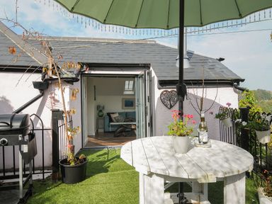 A patio with a round white table and umbrella next to a house with open doors showing a living room at Moons Cottage in Fowey