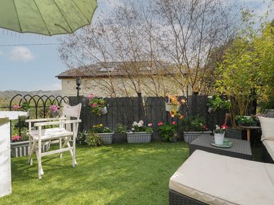An outdoor patio area with artificial grass a white chair a wicker table with potted plants and a black privacy screen with flowers at Moons Cottage in Fowey