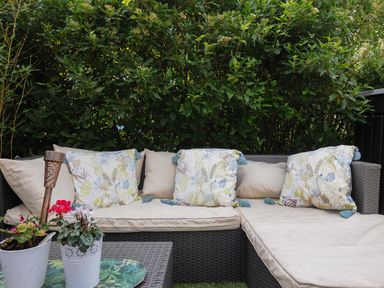An outdoor wicker sofa with cushions on a patio next to green bushes at Moon's Cottage in Fowey