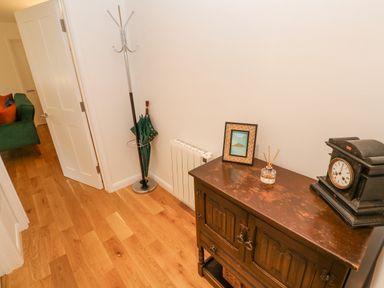 A hallway with a wooden cabinet holding a clock a framed picture and a diffuser and a coat stand with an umbrella near an open door and a green sofa at James Place in Falmouth
