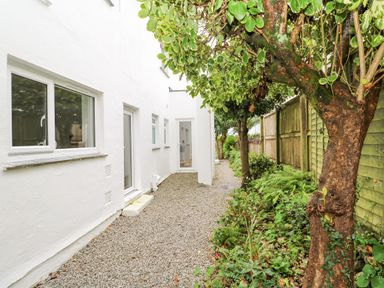 A gravel pathway alongside a white building with windows and doors and a wooden fence with trees and plants at James Place in Falmouth