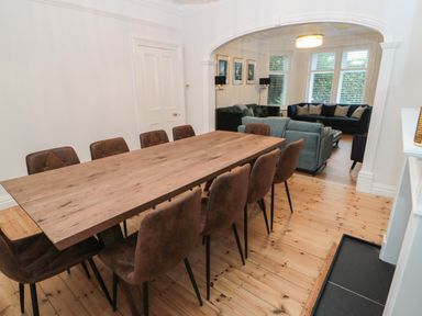 A dining room with a wooden table and brown chairs next to a living room with sofas and large windows at St Aidans in Alnwick