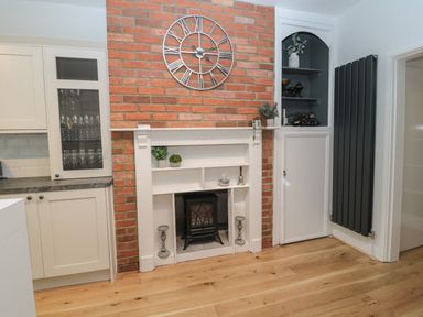 A kitchen area with white cabinets glass door cupboards a brick wall with a metal clock white painted built in shelves and a black heater at St Aidans in Alnwick
