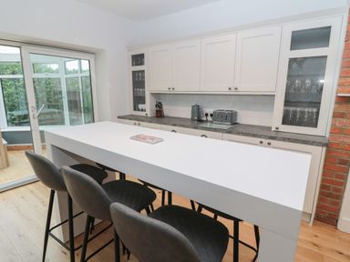 A kitchen with a long white island table and four black chairs next to white cabinets and glass doors at St Aidans in Alnwick