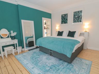 A bedroom with a double bed teal blanket and pillows a large mirror white dressing table and teal accent wall at St Aidans in Alnwick