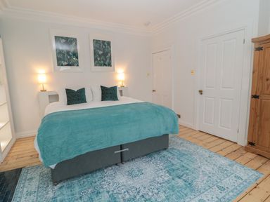A bedroom with a double bed covered with teal bedding and two pillows two lamps on bedside tables two framed pictures on the wall a wooden wardrobe and two white doors at St Aidans in Alnwick