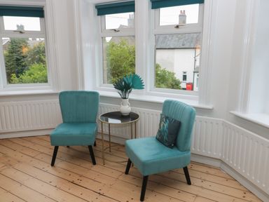 A sitting area with two teal chairs and a small round table with a white vase and teal leaves in front of windows with white frames at St Aidans in Alnwick
