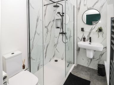 A bathroom with a shower enclosure a toilet a wall-mounted sink a round mirror and a towel heater at St Aidans in Alnwick