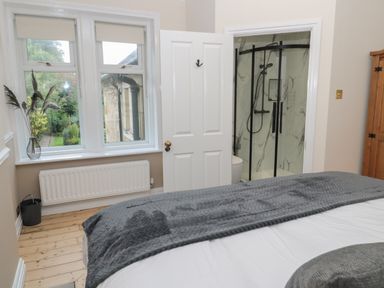 A bedroom with a grey blanket on the bed a window with blinds and a vase with plants and a door leading to a bathroom with a glass shower at St Aidans in Alnwick