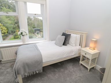 A bedroom with a single bed with pillows and a gray blanket a bedside table with a lamp and a window with a vase of flowers at St Aidans in Alnwick