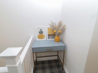 A blue console table with a yellow vase and two framed artworks in a corner by a staircase railing at St Aidans in Alnwick