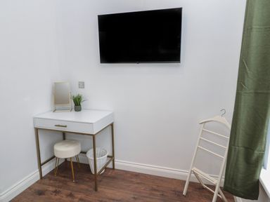 A small white desk with a mirror and plant a round stool a white basket a wall mounted television and a clothes valet with a green curtain in a room at St Aidans in Alnwick