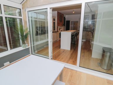 A kitchen with a white island and stools viewed through sliding glass doors from a room with a white table and a potted plant at St Aidans in Alnwick