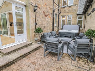 An outdoor patio with grey cushioned chairs around a glass table a hot tub and two potted plants at St Aidans in Alnwick